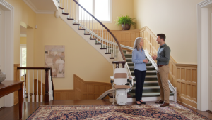 Two people stand and talk beside a stairlift, highlighting thoughtful Mobility Adaptions in a well-lit home with beige walls, wooden accents, and a large area rug.