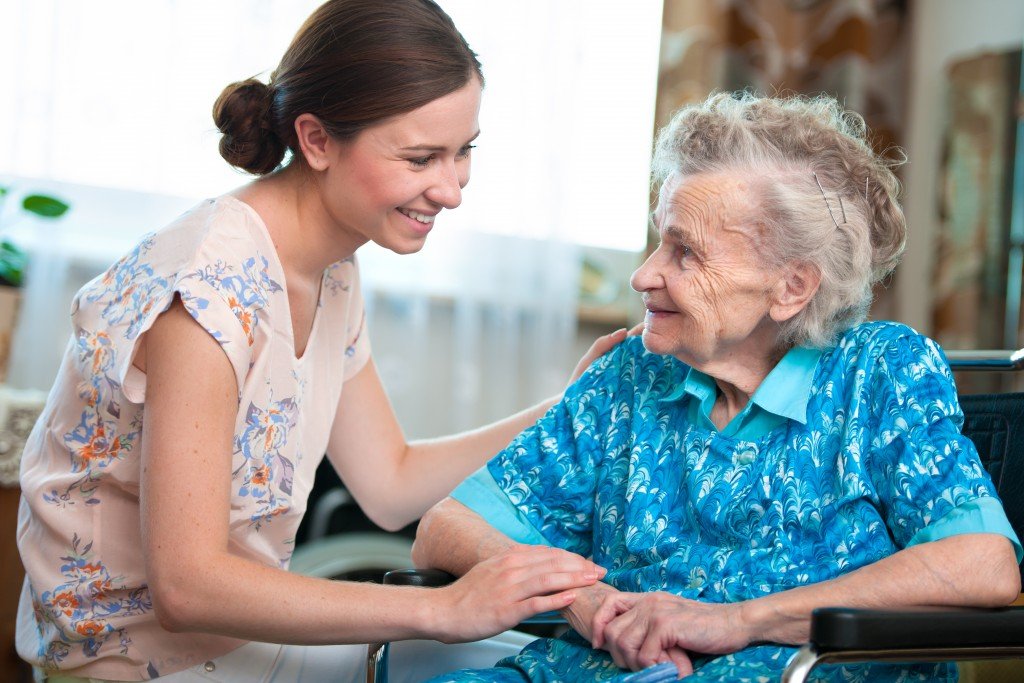 A young woman smiles while holding hands with an elderly woman seated in a wheelchair, a piece of essential mobility equipment. They are indoors, with light streaming through a window in the background.