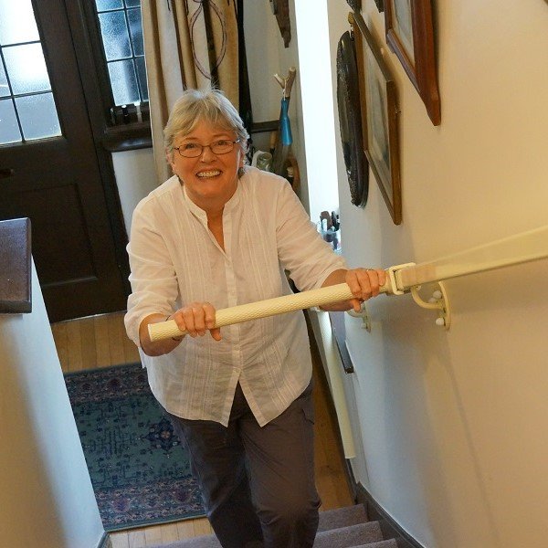 An older woman smiling while using a stairlift in her well-lit home, wearing a white shirt and glasses.