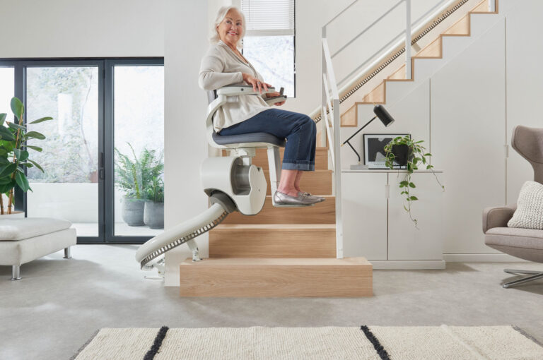 An older woman is using a stairlift in a modern home living room, ascending beside a white staircase. She is smiling and holding a remote control device.