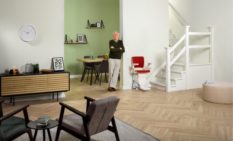 A man stands in a modern living room with light green and white walls, a wooden floor, a small dining area, a staircase equipped with stairlifts, and minimalist decor items.