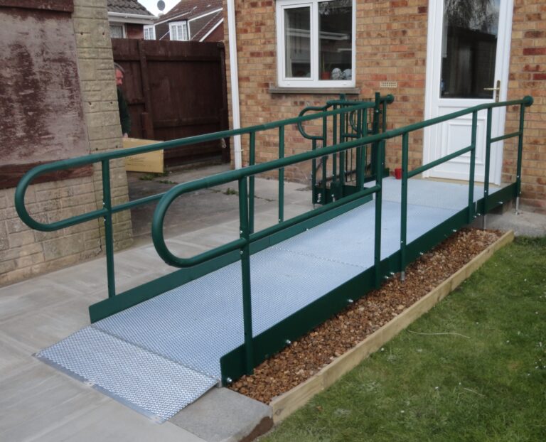 A green metal wheelchair ramp with handrails, designed for accessibility, leads to a white door on a brick building. The ramp is supported by a sturdy base of concrete and gravel.