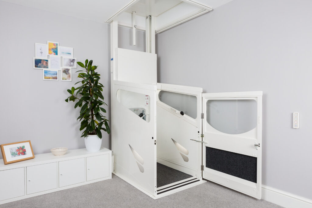 A white two-level home lift, unlike other lifts, is elegantly installed in a gray room next to a potted plant and a white cabinet adorned with framed pictures.