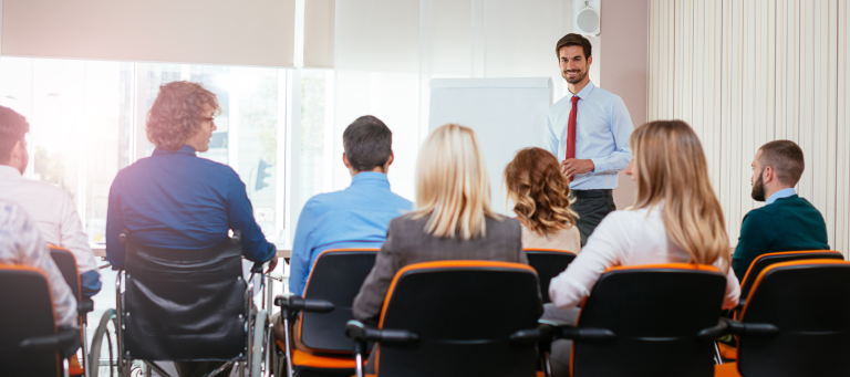 A man in a shirt and tie stands and presents to a group of people sitting on orange chairs in a conference room, emphasizing the importance of workplace accessibility.