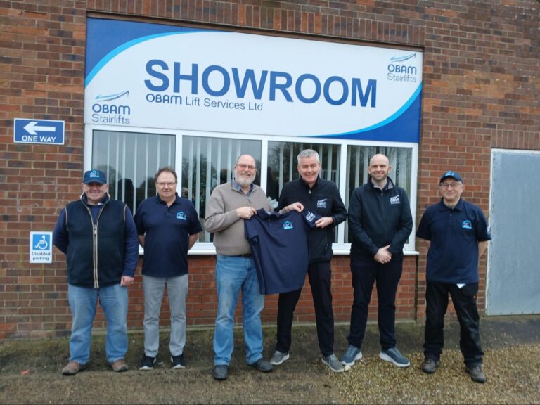 Six men gather proudly in front of the OBAM Lift Services Ltd showroom sign, a testament to community support. One man holds up a jacket, perhaps a memento from the Shed Club, illustrating their unity and shared purpose.