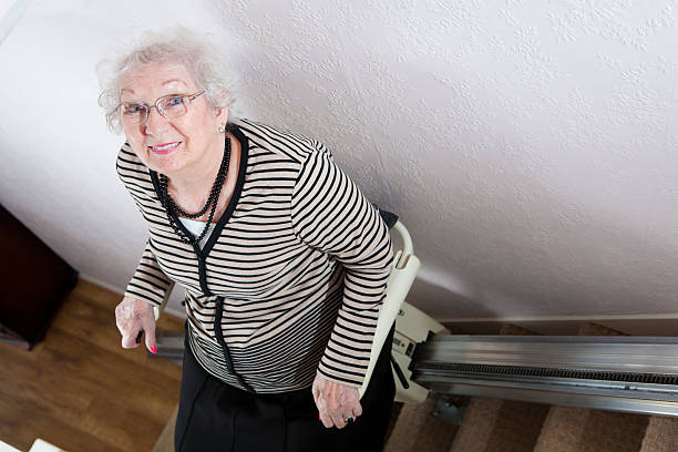 Elderly woman with glasses smiles while sitting on a stairlift at the top of a staircase in a home, highlighting the benefits of stairlift financial support for improved home mobility UK.