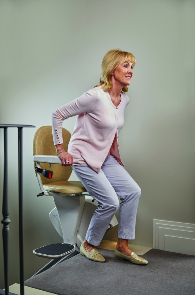 An older woman stands up from a Synergy Straight Stairlift at the top of a staircase in a comfortable home setting.