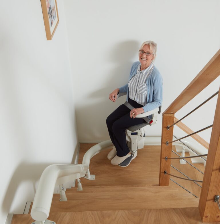 An older woman sits on a Platinum Ergo stairlift at the top of a wooden staircase, smiling at the camera.