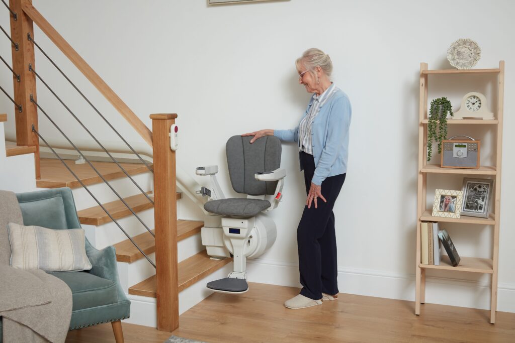 An older woman stands by an Ultimate stairlifts curved stairlift at the bottom of a staircase in a living room, preparing to use it.
