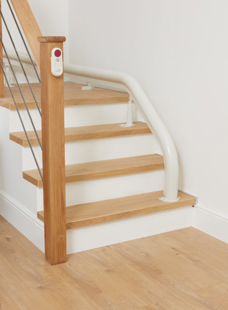 A wooden staircase with a light-colored handrail and a stairlift rail, compatible with curved stairlifts like the Platinum Ergo, set against a white wall and light wood floor.