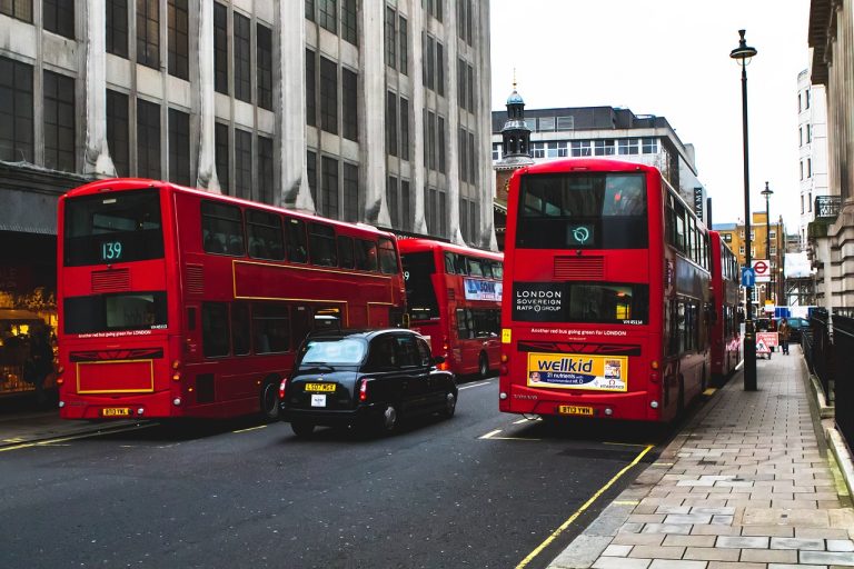 Two red double-decker buses and a black taxi navigate a city street lined with tall buildings in London, where many residents enjoy mobility discounts once they reach the bus pass age UK eligibility.