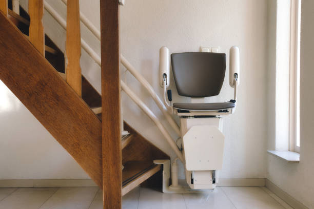A stair lift chair is installed beside a wooden staircase in a well-lit interior space, positioned at the bottom of the stairs near a window. For more details, consult our Complete Stairlift Guide.