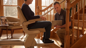 A technician with a tablet consults with a person seated on a stairlift at the base of a wooden staircase inside a home, conducting stairlift assessments as part of the home survey process.