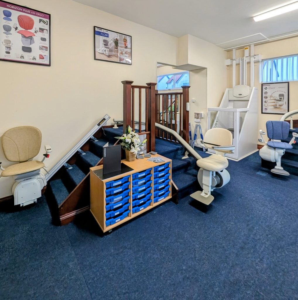Showroom with multiple stairlift models installed on sample staircases, informational posters on walls, and a cabinet with blue drawers in the center of the room.