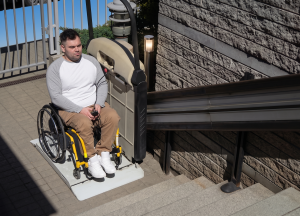 A man in a wheelchair uses a platform lift to access a staircase next to a brick wall outdoors, demonstrating stairlift safety and accessibility.