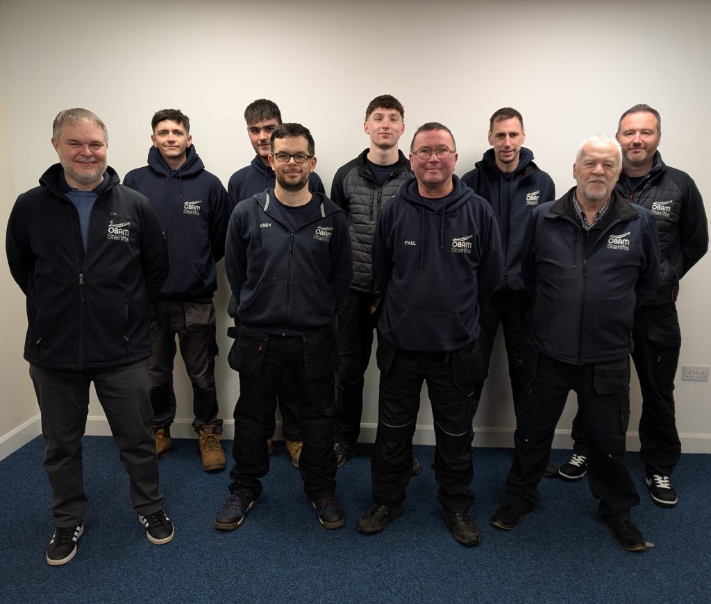 Eight men stand indoors against a plain wall, all wearing dark uniforms with company logos, posing for a group photo on a blue carpeted floor, dedicated to stairlift personalisation and transparent stairlift pricing.