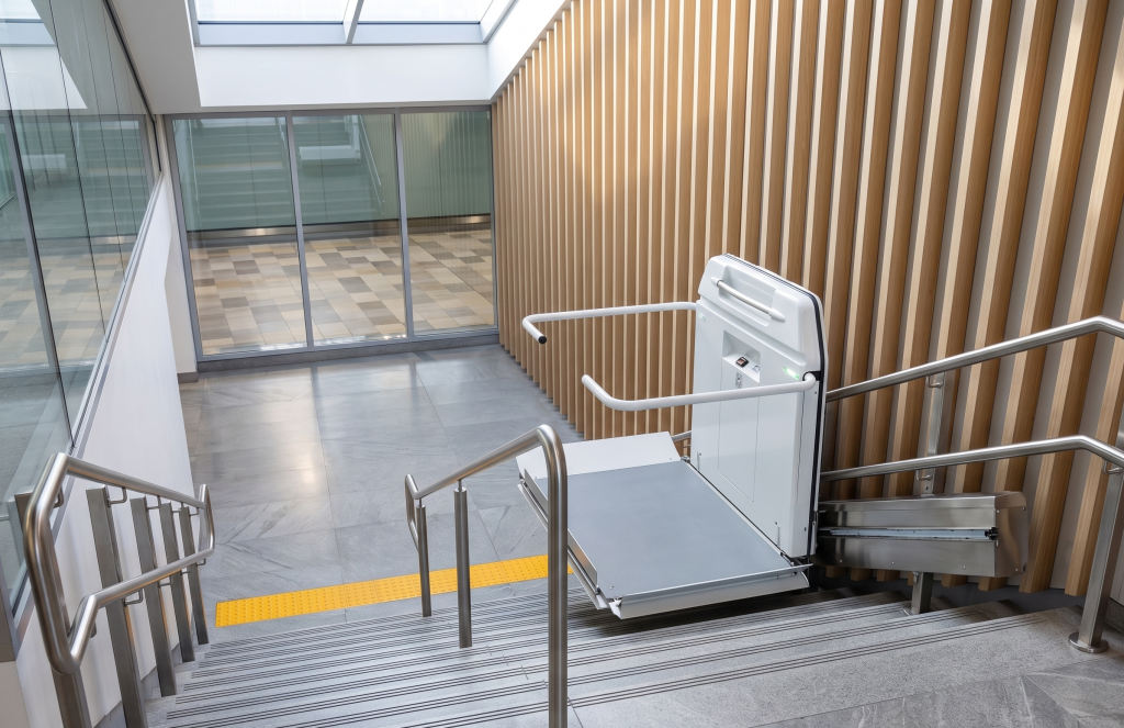 A wheelchair lift installed on a staircase in a modern building, with metal railings and a tactile yellow strip at the top of the stairs—an excellent example for those doing a stairlift comparison or researching the best stairlifts.