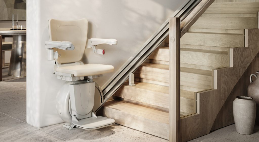 A Savaria K2 straight stairlift is installed alongside a wooden staircase in a modern home, with neutral-coloured walls and pottery decor nearby.