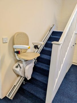 A stairlift installed along a blue-carpeted staircase with white railings in an indoor setting.