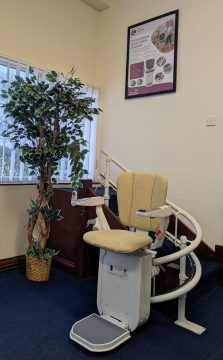 A stairlift with a cream-colored seat is installed on a curved staircase in a carpeted room next to a potted plant and a window. A poster is mounted on the wall above.