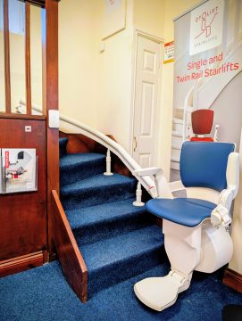A stairlift installed on a short flight of carpeted stairs in an indoor showroom, with a banner and instruction display on the wall.