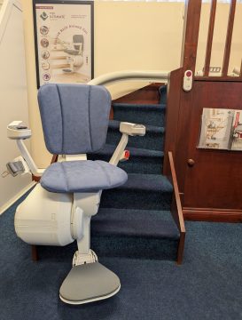 A stairlift with a blue cushioned seat is installed on a carpeted staircase; promotional posters and a control panel are visible on the wall.