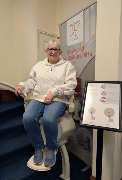An older woman sits and smiles on a stairlift in a showroom next to a promotional banner and a product information board for Otolift stairlifts.